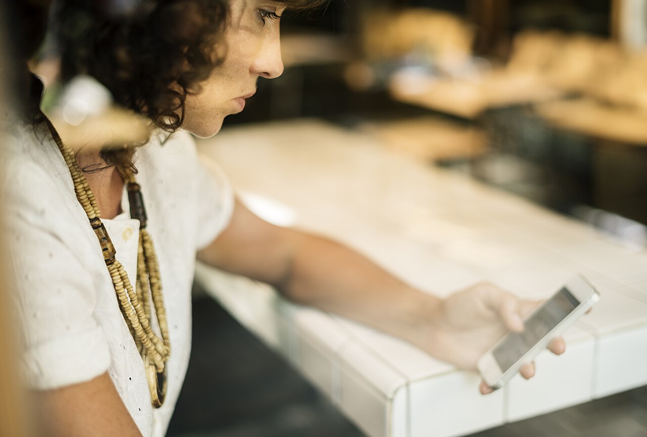 A woman with curly hair and a beaded necklace looks at her smartphone beside a white tiled counter.